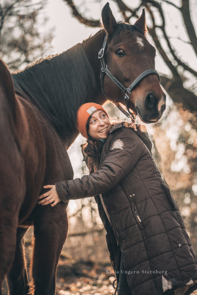 Bild von einer Frau und einem braunen Hannoveraner Pferd in einem Wald.